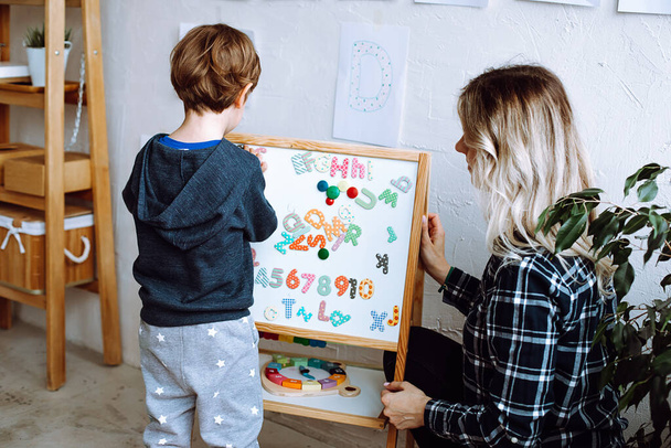 Kindergartenkinder stehen mit Rücken und lernen englische Buchstaben, Alphabet und Zahlen auf einer Magnettafel. Erzieher sitzen neben Jungen im Spielzimmer und kontrollieren Bewegung. Lesen und Zählen, Lernen in der Kita.  - Foto, Bild