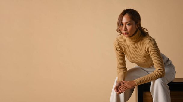 Впевнена молода жінка в модному светрі сидить на бежевому фоні. Осіння концепція моди та краси
. - Фото, зображення