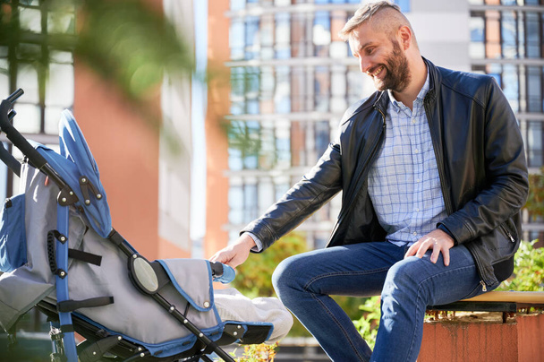 Father entertaining little son in stroller, while walking on street. Young bearded man making newborn baby laugh, while sitting on bench at modern courtyard of city residential high-rise buildings. - Photo, Image