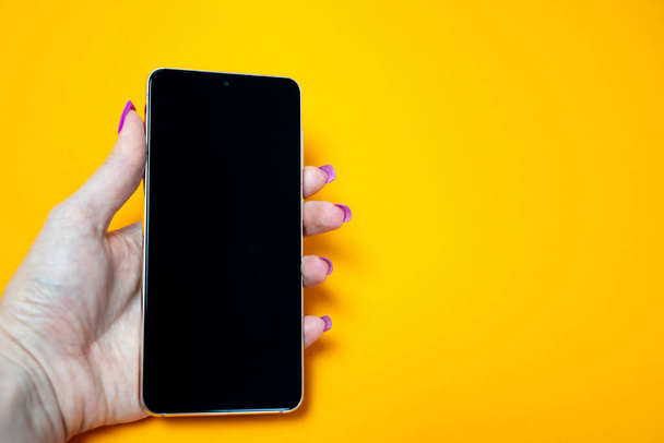 женщина рука с ногтями маникюр держа смартфон с чистым черным экраном на желтом фоне, место, пространство для текста, вставка изображения, современные технологии, подключение к Интернету, Wi-Fi, обмен сообщениями  - Фото, изображение