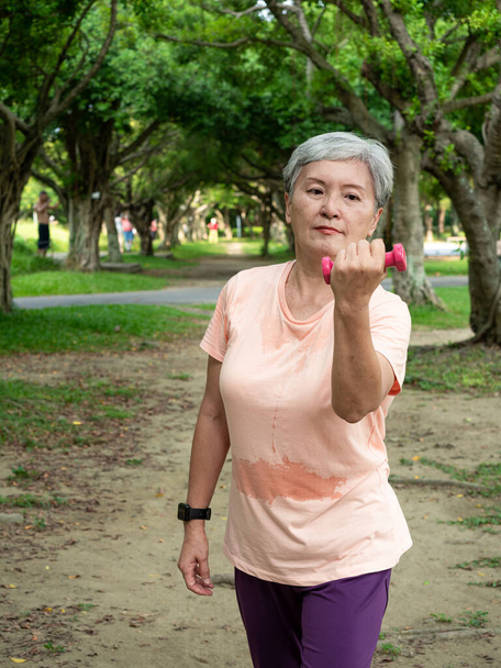 Портрет зрелой азиатки 60-х годов в спортивной одежде, занимающейся спортивной тренировкой рук и поднимающей гантели с расслаблением для здорового в парке на открытом воздухе. - Фото, изображение