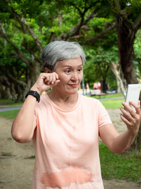 高齢のアジア系の女性の肖像60代の笑顔立っていると公園でスマートフォンを使用して眼精を持っています. - 写真・画像