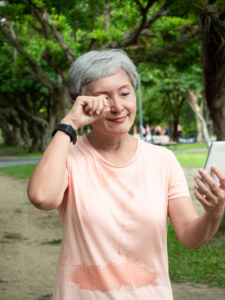 Porträt einer älteren, erwachsenen asiatischen Frau um die 60, die im Stehen lächelt und ihr Smartphone im Park benutzt. - Foto, Bild