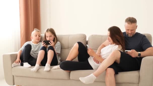 Amerikalı aile, aileler ve küçük çocuklar gülüyor, akıllı telefon cihazları kullanıyorlar, evdeki kanepede birlikte dinleniyorlar, akıllı telefon ekranlarına bakıyorlar, mobil uygulama kullanıyorlar. - Video, Çekim