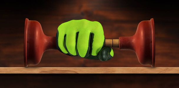 Hand with green and black protective work glove, holding a red rubber plunger with wooden handle, on a wooden workbench with copy space. - Photo, Image