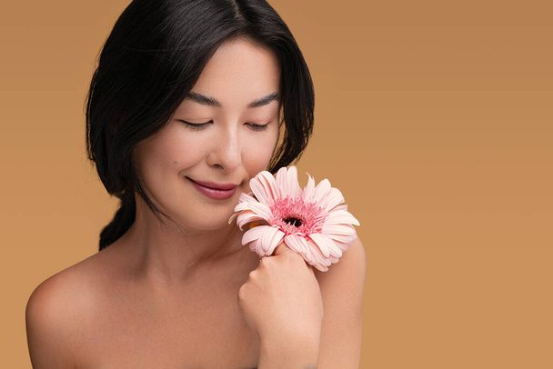 Beautiful young ethnic female model with pure skin performing perfect cosmetic skincare holding blooming flower on brown background - Foto, immagini