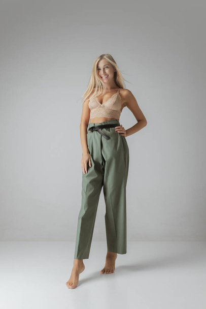 Attractive young woman with long straight blonde hair and natural makeup wearing crop top and casual trousers posing over grey background. Natural beauty, casualoutfit, inspiration, fashion and ad - Photo, Image
