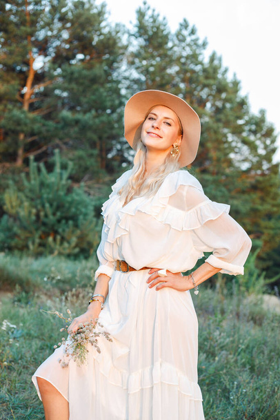 Retrato de una hermosa mujer sonriente con el pelo largo y rubio con vestido blanco, sombrero beige floppy, parada afuera frente a los árboles en el parque forestal, sosteniendo ramo de flores silvestres en verano. Vertical. - Foto, imagen