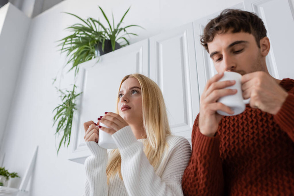 low angle view of young woman in knitted sweater holding cup of tea near blurred boyfriend  - 写真・画像