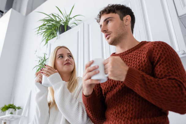 low angle view of young woman in knitted sweater holding cup of tea while looking at boyfriend  - Photo, Image