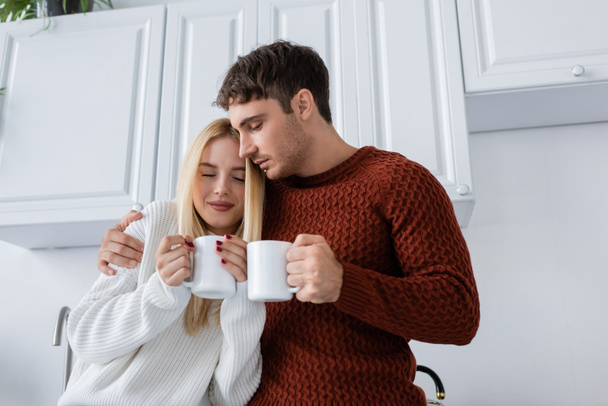young couple in knitted sweaters hugging and holding cups with tea while warming up in winter - Photo, image