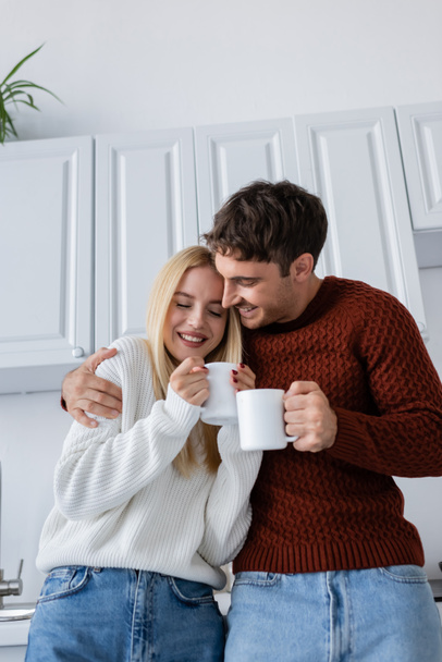 счастливая молодая пара в вязаных свитерах обнимает и держит чашки с чаем, согреваясь зимой - Фото, изображение