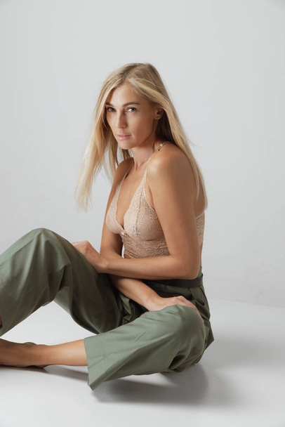 Attractive young woman with long straight blonde hair and natural makeup wearing crop top and casual trousers posing over grey background. Natural beauty, casualoutfit, inspiration, fashion and ad - 写真・画像