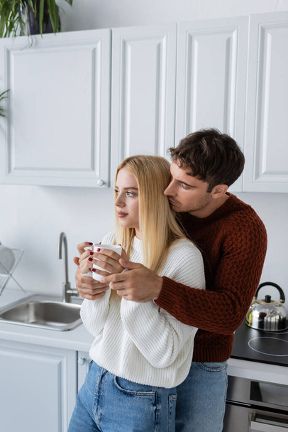 young man in knitted sweater hugging blonde woman with cup of tea during winter - Foto, imagen