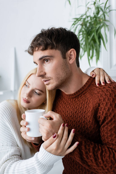 blonde woman hugging boyfriend in red sweater with cup of tea  - Фото, изображение
