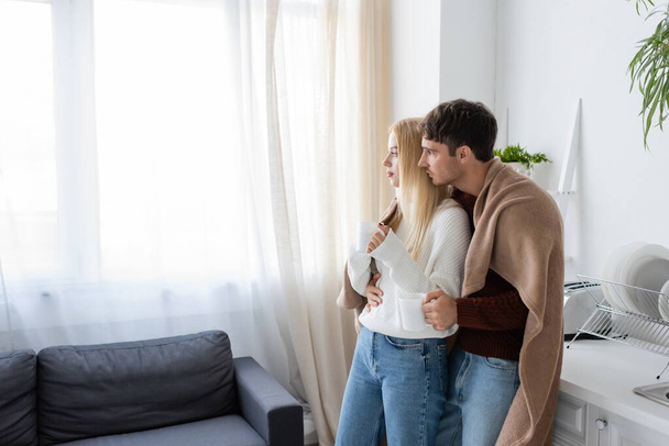 young couple covered in blanket hugging while holding cups of tea at home - Photo, Image