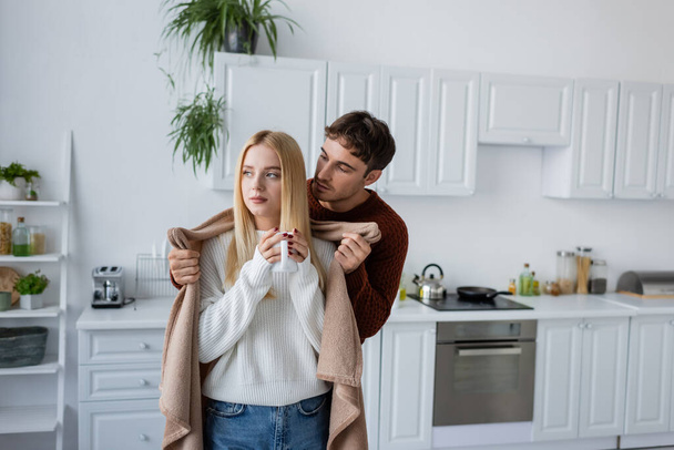 caring man holding blanket near girlfriend in sweater with cup of tea - Foto, afbeelding
