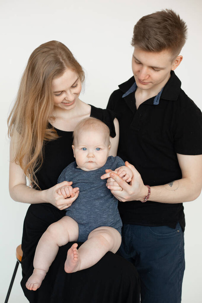 Fiatal, mosolygós, vonzó család portréja sötét ruhában, kis szürke szemű kisbabával a fehér háttéren. A szülők fogják a gyerek kezét. Család, kapcsolat, szórakozás. - Fotó, kép