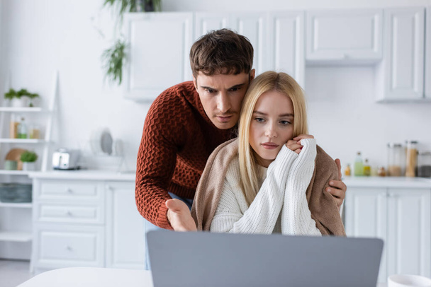 confused man in sweater and blonde woman looking at laptop while working from home - Fotó, kép