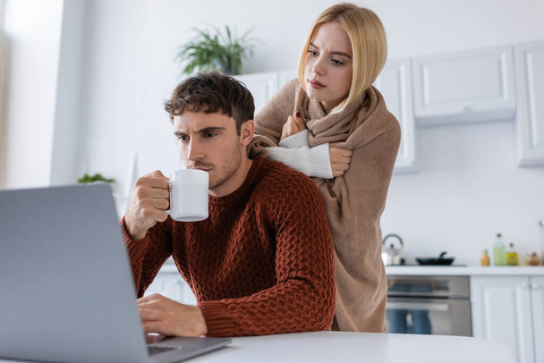 mujer rubia cubierta de manta de pie detrás de novio beber té mientras trabaja desde casa  - Foto, Imagen