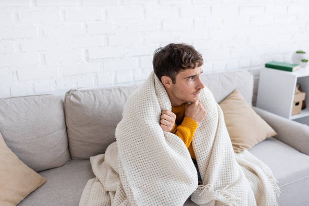 junger Mann in Decke gehüllt sitzt auf Sofa und wird warm im Wohnzimmer  - Foto, Bild
