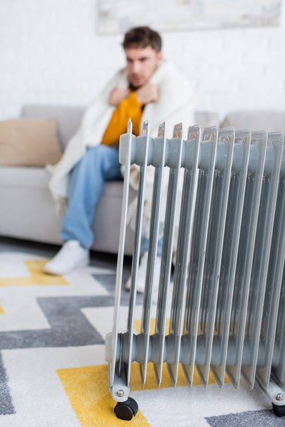 radiador calentador cerca borrosa joven cubierto de manta sentado en el sofá en la sala de estar  - Foto, Imagen