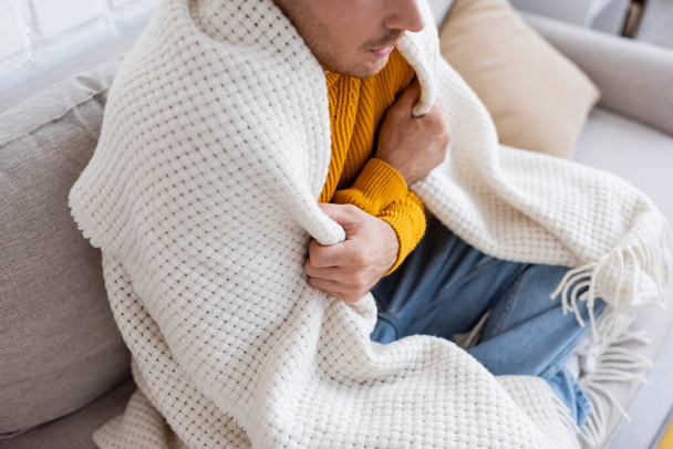high angle view of young man covered in blanket sitting on sofa and getting warm in living room  - Photo, Image