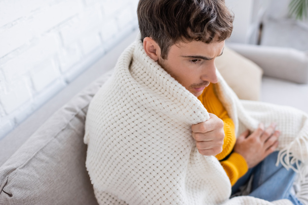high angle view of young man covered in blanket sitting on sofa and getting warm at home - Foto, imagen