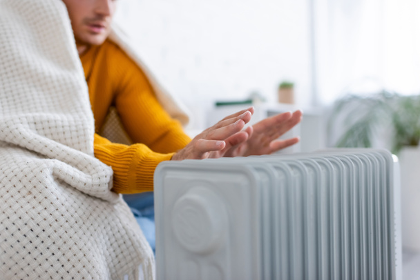 visão parcial do jovem coberto de cobertor sentado no sofá e aquecimento perto do aquecedor do radiador - Foto, Imagem