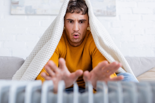Emotionaler junger Mann in Decke gehüllt wärmt Hände neben Heizkörper - Foto, Bild