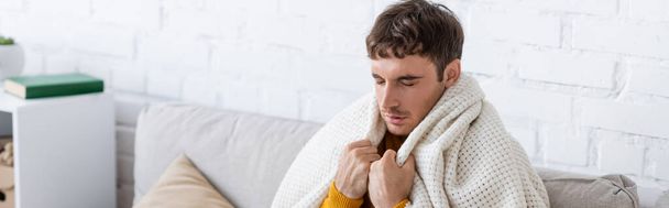 young man with closed eyes holding blanket while sitting on sofa and getting warm at home, banner - Foto, immagini