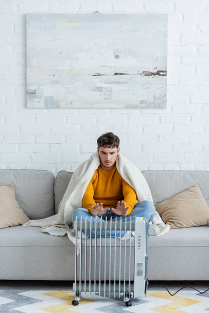 jovem coberto de cobertor sentado no sofá e aquecendo as mãos perto do aquecedor radiador moderno  - Foto, Imagem