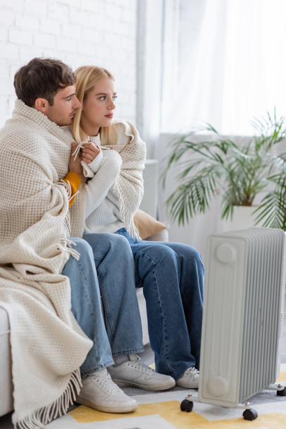 young couple covered in warm blanket sitting on couch near radiator heater at home - Photo, Image