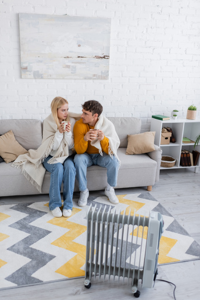 jovem casal coberto de cobertor sentado com xícaras de chá perto aquecedor radiador - Foto, Imagem