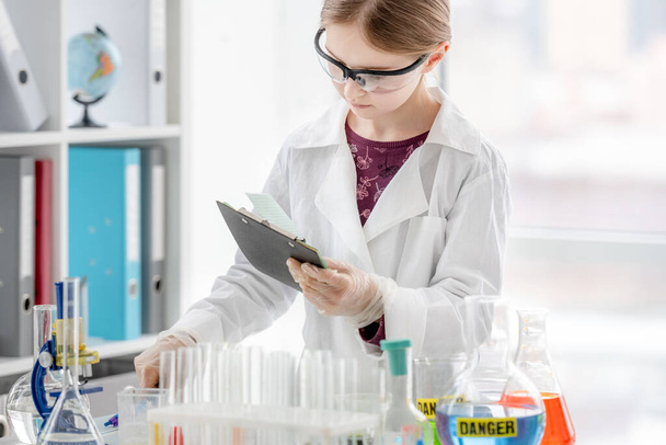 Meisje tijdens wetenschappelijk chemisch experiment maakt notities dicht bij tafel met buizen. Schoolmeisje met chemische apparatuur op school - Foto, afbeelding