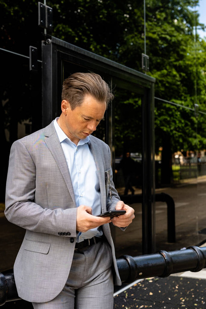 Un bell'uomo d'affari con un completo grigio in piedi fuori dal suo ufficio mentre usa il suo cellulare. Pranzo di lavoro - Foto, immagini