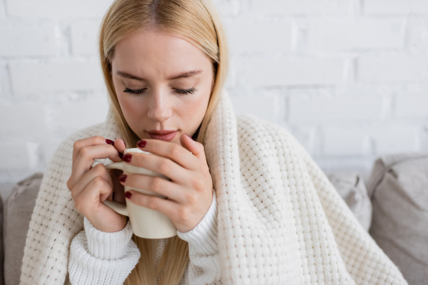 ξανθιά γυναίκα σε πλεκτό πουλόβερ κάθεται καλυμμένη με κουβέρτα με φλιτζάνι τσάι στο σαλόνι  - Φωτογραφία, εικόνα