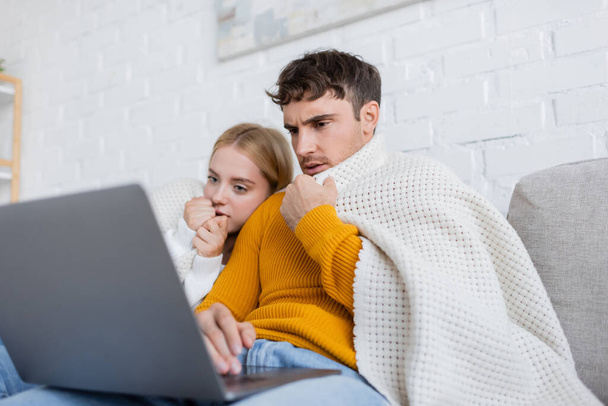 couple covered in blanket watching scary movie on laptop in living room  - Zdjęcie, obraz