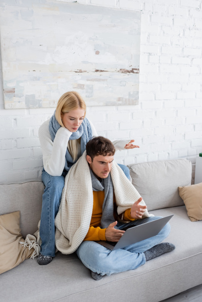 ontevreden blonde vrouw in trui en sjaal zitten in de buurt freelancer en gebaren terwijl het kijken naar laptop  - Foto, afbeelding