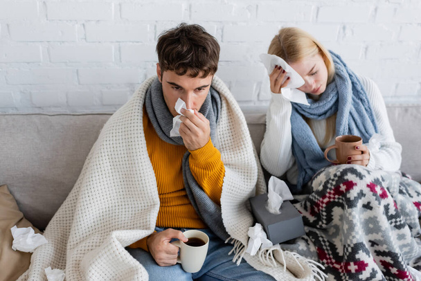 sick couple covered in blankets holding cups of tea and tissues while sitting on sofa - Foto, immagini
