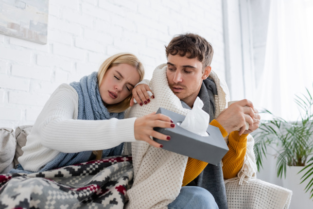 blonde woman in scarf holding tissue box near diseased boyfriend with cup of tea - Photo, Image