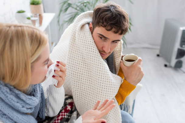 vista de ángulo alto de la mujer rubia enferma en bufanda estornudando en tejido cerca de novio enfermo con taza de té - Foto, imagen