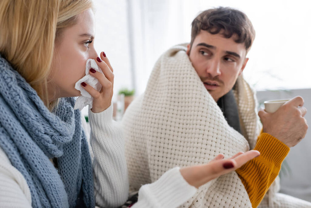 mujer rubia enferma en bufanda estornudando en tejido cerca de novio enfermo con taza de té - Foto, Imagen
