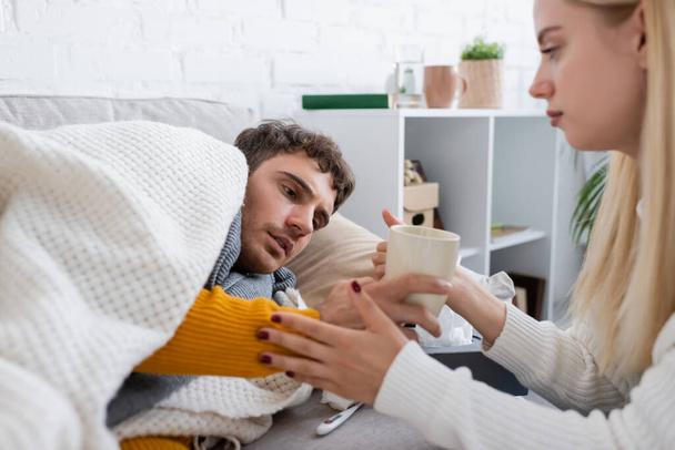 femme attentionnée tenant une tasse de thé près du petit ami malade couché sur le canapé sous la couverture - Photo, image