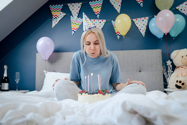 Happy woman in pajama and party cap blowing out candles on birthday cake on the bed in decorated bedroom. Make a wish process. Morning surprise. Happy birthday concept. Selective focus - Foto, Imagem