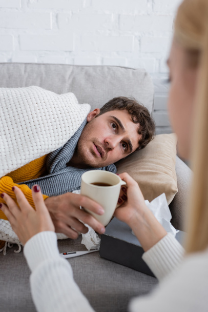 Заботливая девушка с чашкой чая возле больного парня, лежащего на диване под одеялом - Фото, изображение