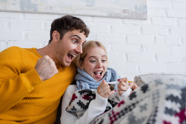 excited man and sick blonde woman looking at digital thermometer  - Фото, изображение