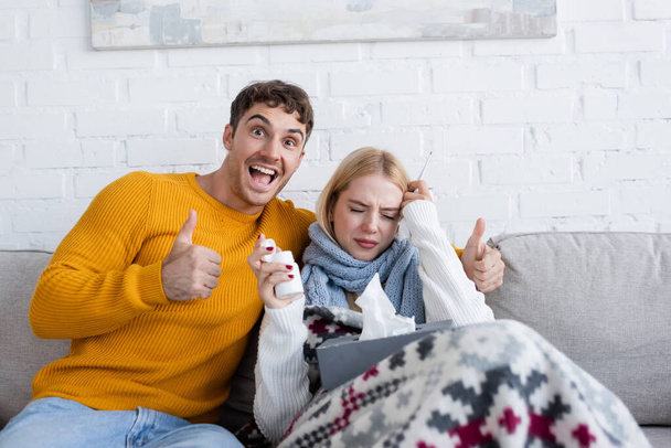 cheerful man showing thumb up near sick blonde woman with tissue box  sitting on sofa - Fotoğraf, Görsel