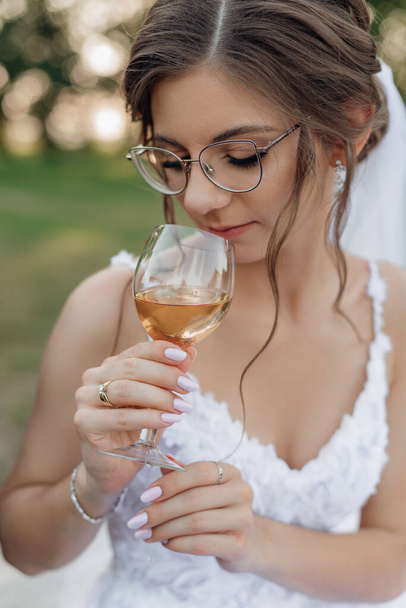 Portrait of thoughtful amazing woman with beautifully coiffured dark hair wearing glasses, white dress, standing outside in park in summer, holding glass of wine, sniffing. Wedding, beauty. Vertical. - Foto, Imagem