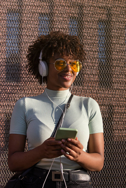 Cool young latin american woman with afro hair and glasses smiling listening to music on smartphone with headphones - Photo, Image